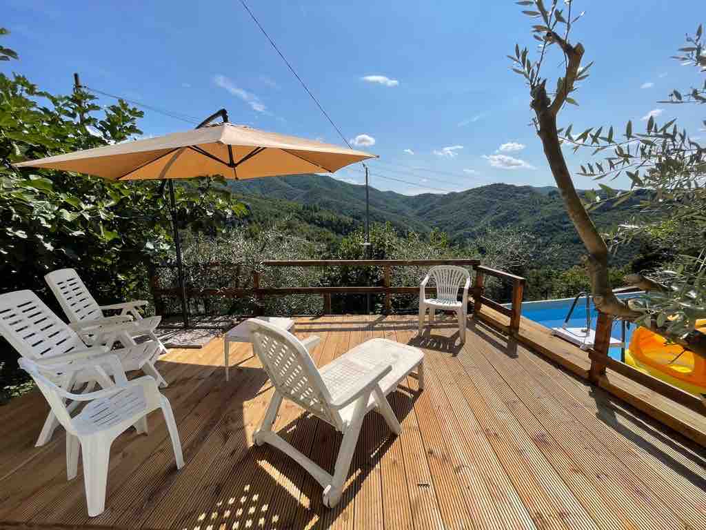 A wooden deck features four white lounge chairs and a large umbrella, providing shade. The surrounding landscape includes lush greenery and distant mountains under a clear blue sky. A swimming pool is visible in the background, enhancing the outdoor relaxation space.