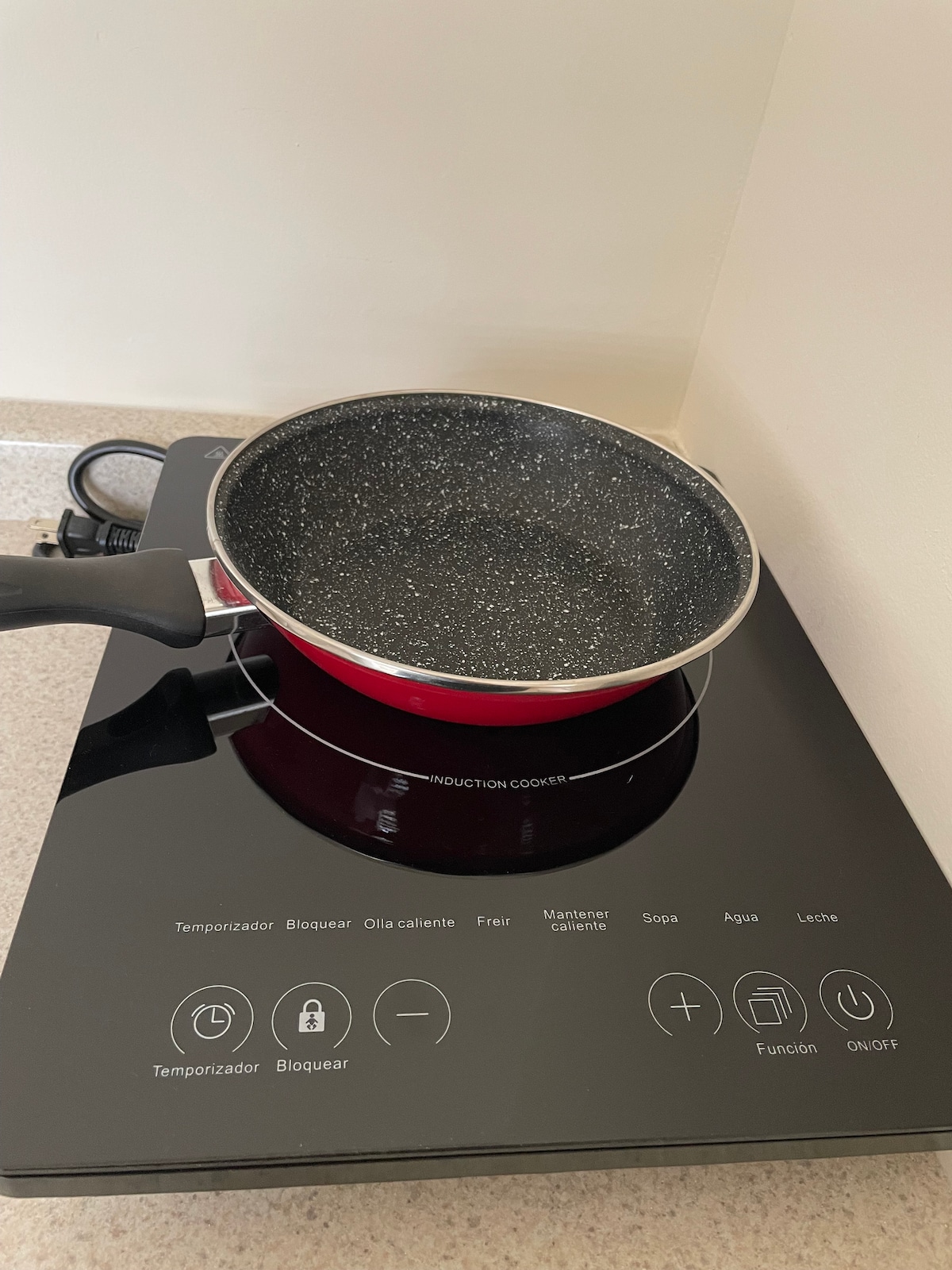 A modern induction cooktop is presented, featuring a sleek black surface with control buttons for temperature and functions. A textured frying pan is placed on the cooktop, noted for its non-stick interior and vibrant red exterior.