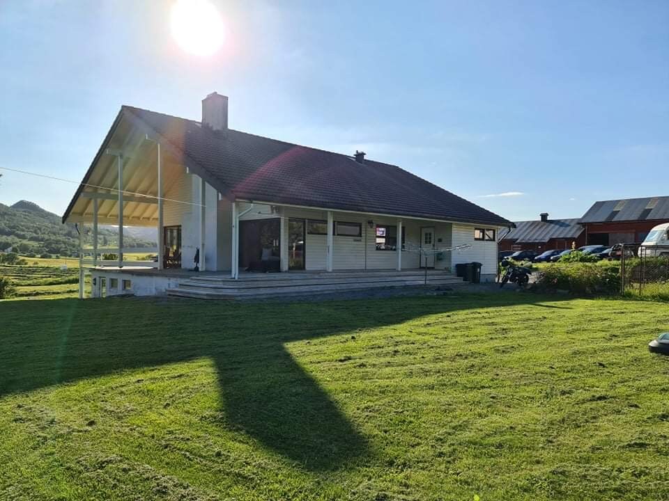 A modern three-story house is set in a rural landscape, characterized by large windows and a sloping roof. A spacious patio is visible, with well-maintained grass surrounding the property. The sun shines brightly, casting a warm glow across the exterior and highlighting the scenic backdrop.