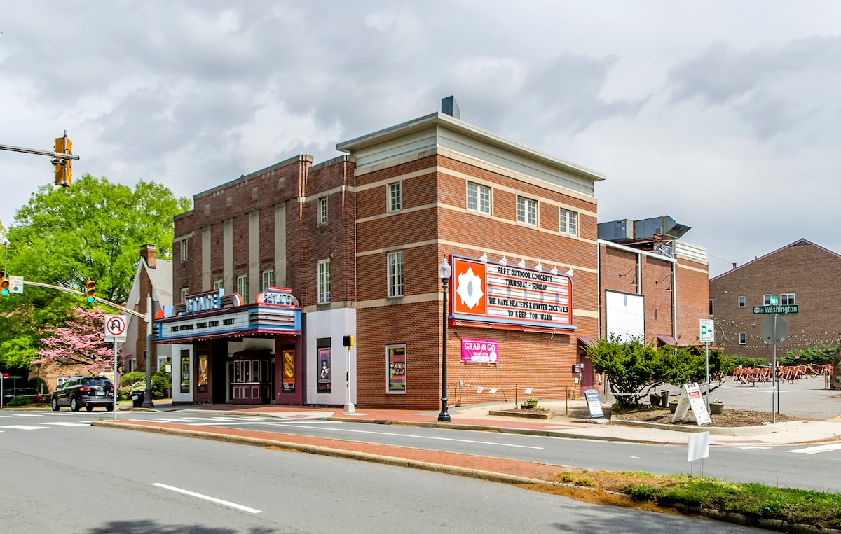 A historic theater building is displayed with its classic brick façade and marquee. Large banners announce upcoming performances, while trees and landscaping provide a natural touch along the street. The scene is well-lit, contributing to the inviting atmosphere of the location.