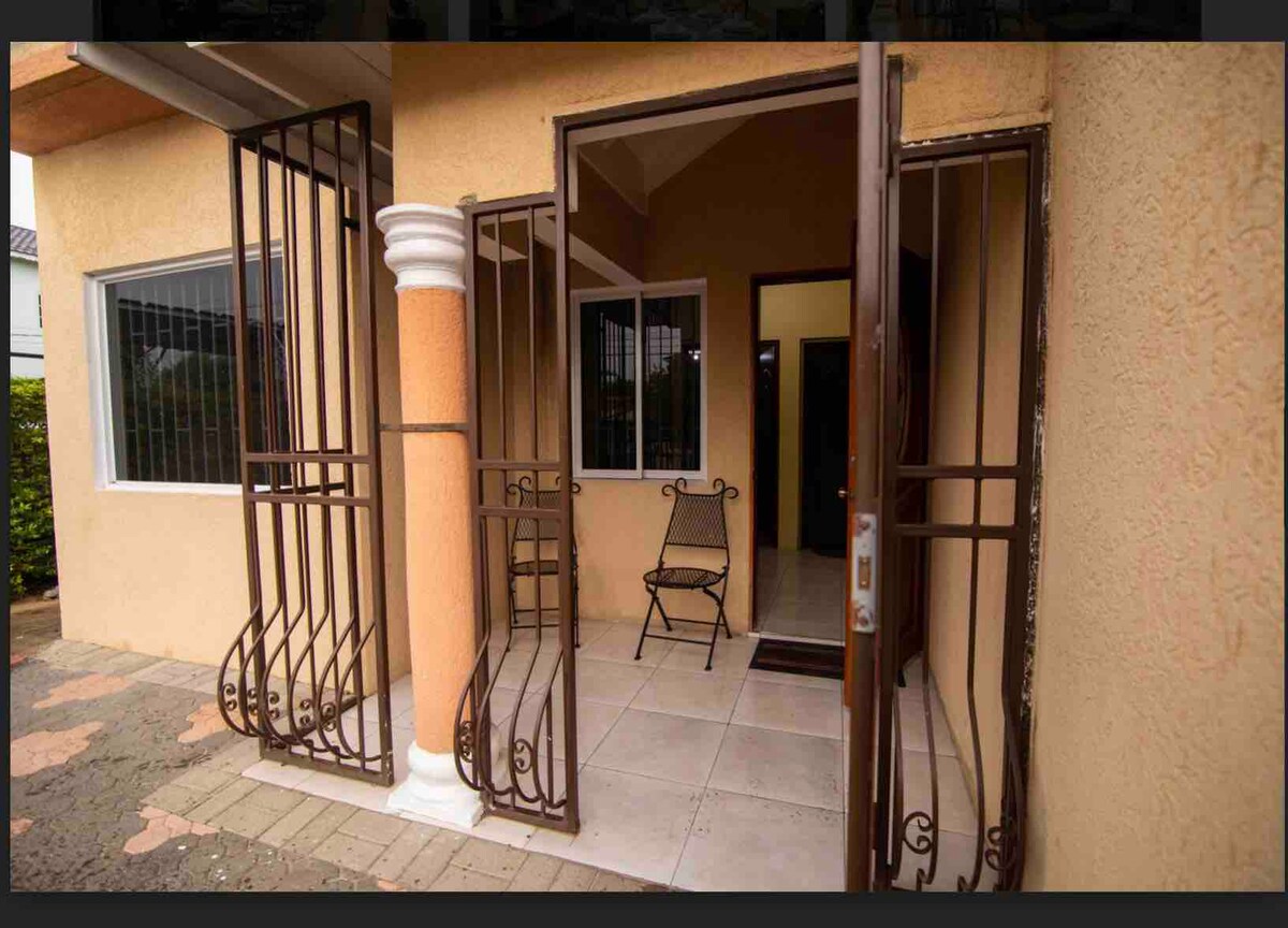 The entrance features a secure gate with decorative ironwork, leading to a spacious foyer area. Two chairs are placed on the patio, offering a comfortable spot to enjoy the outdoors. Large windows allow natural light to fill the space.