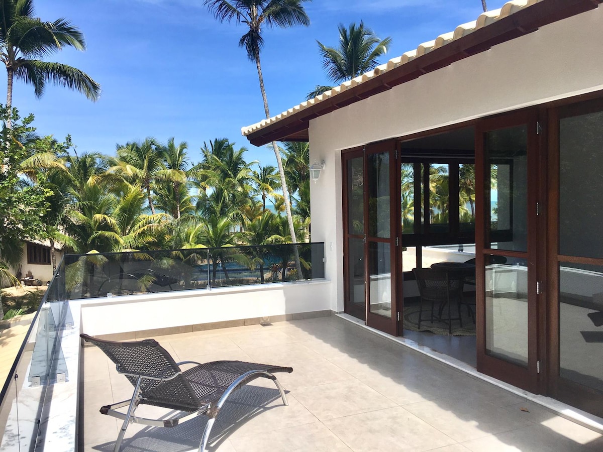 A spacious balcony is highlighted with a sleek lounge chair and surrounded by clear glass railings. Lush palm trees sway gently in the background, complementing the serene coastal view. Natural light floods the area, creating an inviting outdoor space.