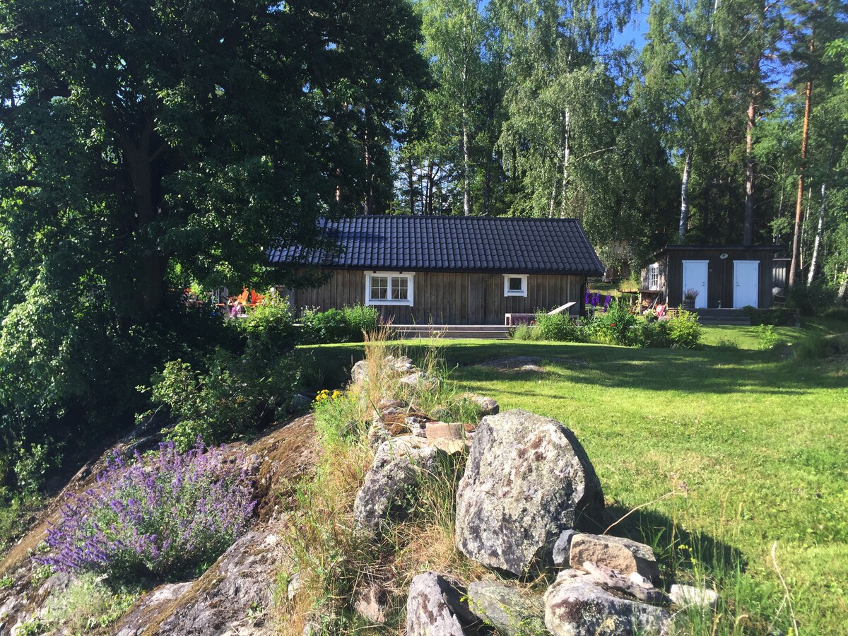 The image shows a charming log cabin surrounded by lush greenery and rocky terrain. A gravel path leads to the entrance, and a secondary structure can be seen nearby. Wildflowers bloom among the rocks, contributing to the serene countryside setting.