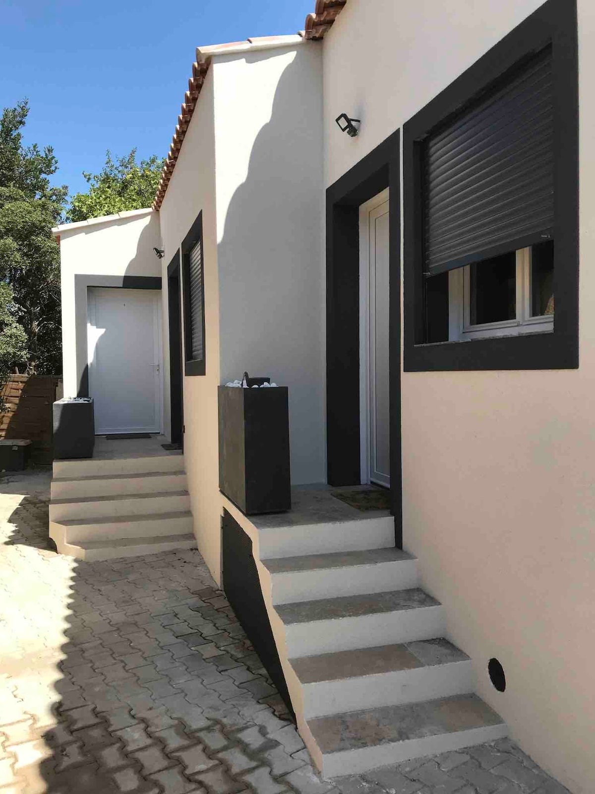 An exterior view of a villa's independent entrance is shown. Two sets of steps lead up to the entrances, which are framed by light-colored walls and dark accents. Windows with shutters provide natural light, while surrounding greenery adds to the peaceful setting.