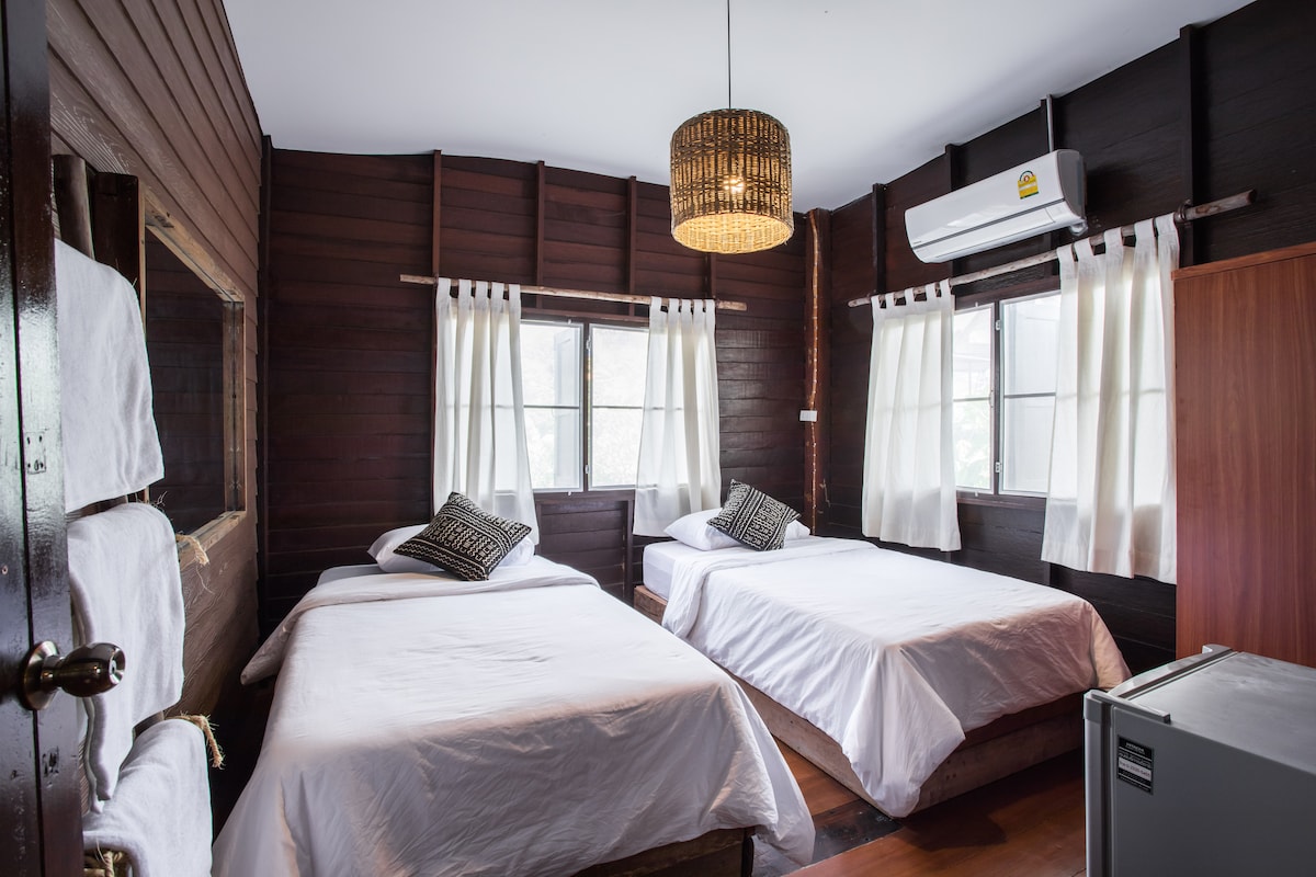 A cozy room features two spacious beds with crisp white linens, accompanied by decorative cushions. Natural light filters through two windows adorned with flowing curtains. A rustic woven pendant light hangs from the ceiling, complementing the wooden wall paneling.