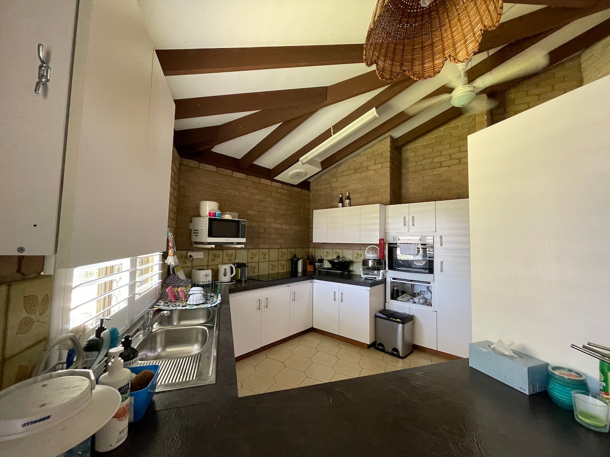The kitchen features a mix of modern appliances and natural elements, with white cabinetry and a textured brick wall. A spacious countertop provides room for meal preparation. Large windows allow natural light to brighten the area, while wooden beams add character to the ceiling.
