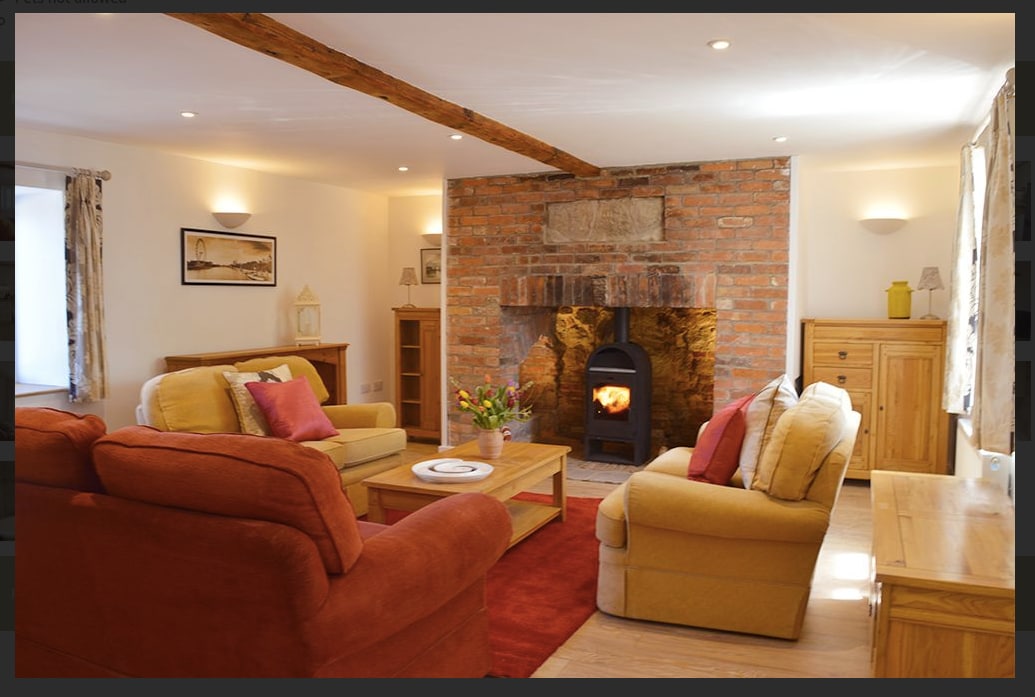 A cozy living area features a brick fireplace with a wood-burning stove, surrounded by comfortable seating arranged around a wooden coffee table. Soft lighting illuminates the room, accentuating original oak beams on the ceiling and creating a welcoming atmosphere.