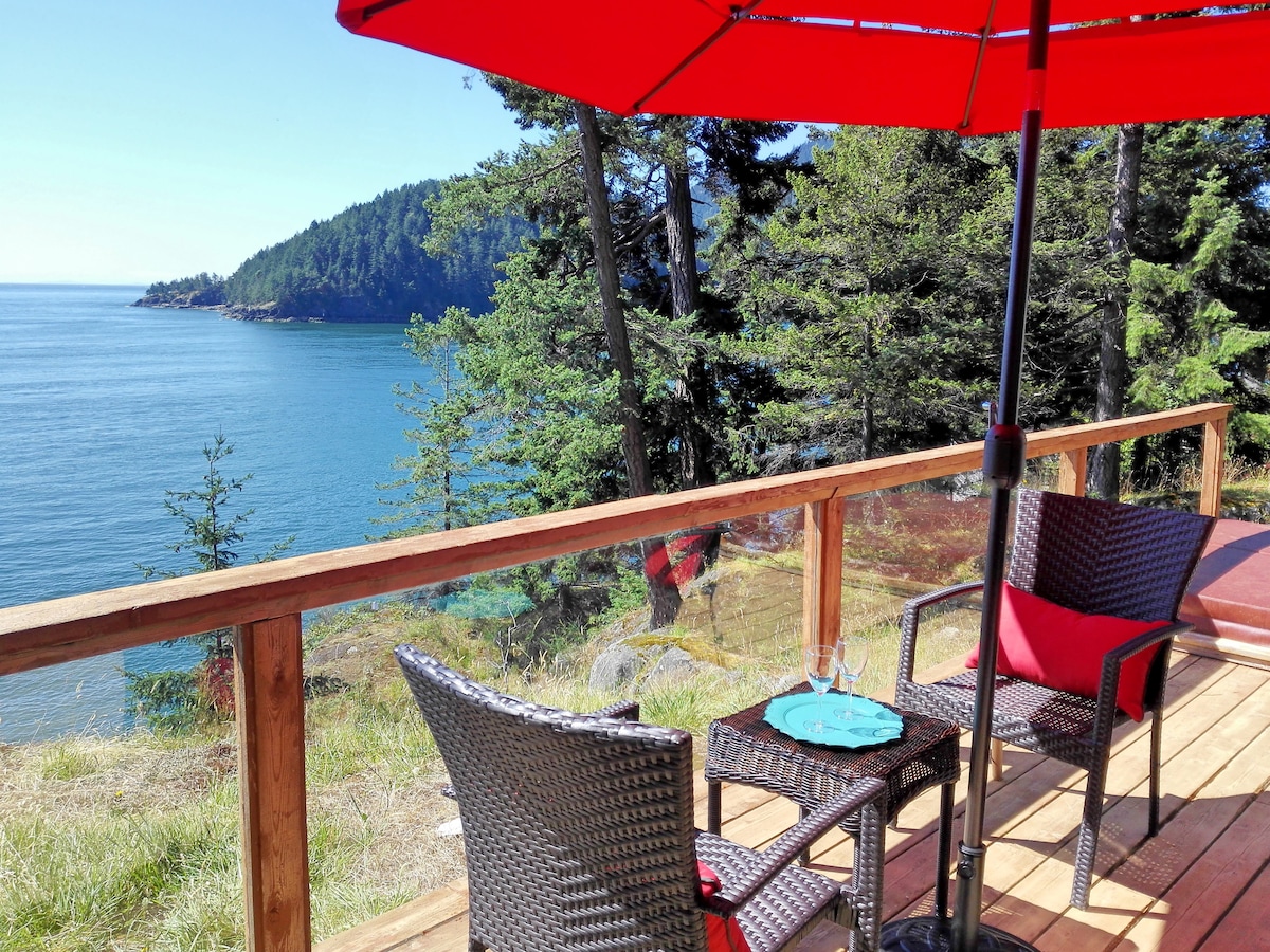 A private deck offers a serene escape with wicker chairs and a small table, set against a backdrop of tranquil water and lush greenery. An umbrella provides shade, while sweeping views of the coastline and distant hills are visible beyond.