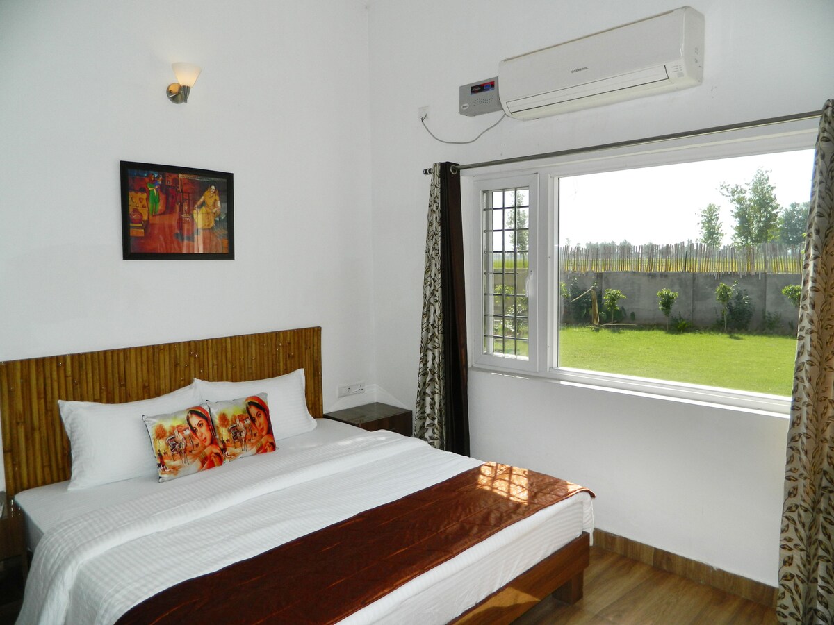 A well-furnished room features a spacious bed with crisp linens and decorative pillows. Natural light enters through a large window overlooking a grassy area. A piece of artwork adorns the wall, while a cooling unit is mounted above for comfort.