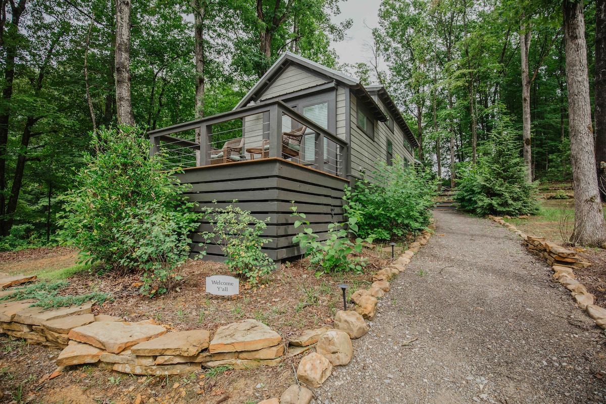A tiny home is nestled among trees, featuring a covered front deck with two chairs and surrounded by greenery. A gravel path leads to the entrance, and natural stone landscape borders the yard, enhancing the tranquil setting.