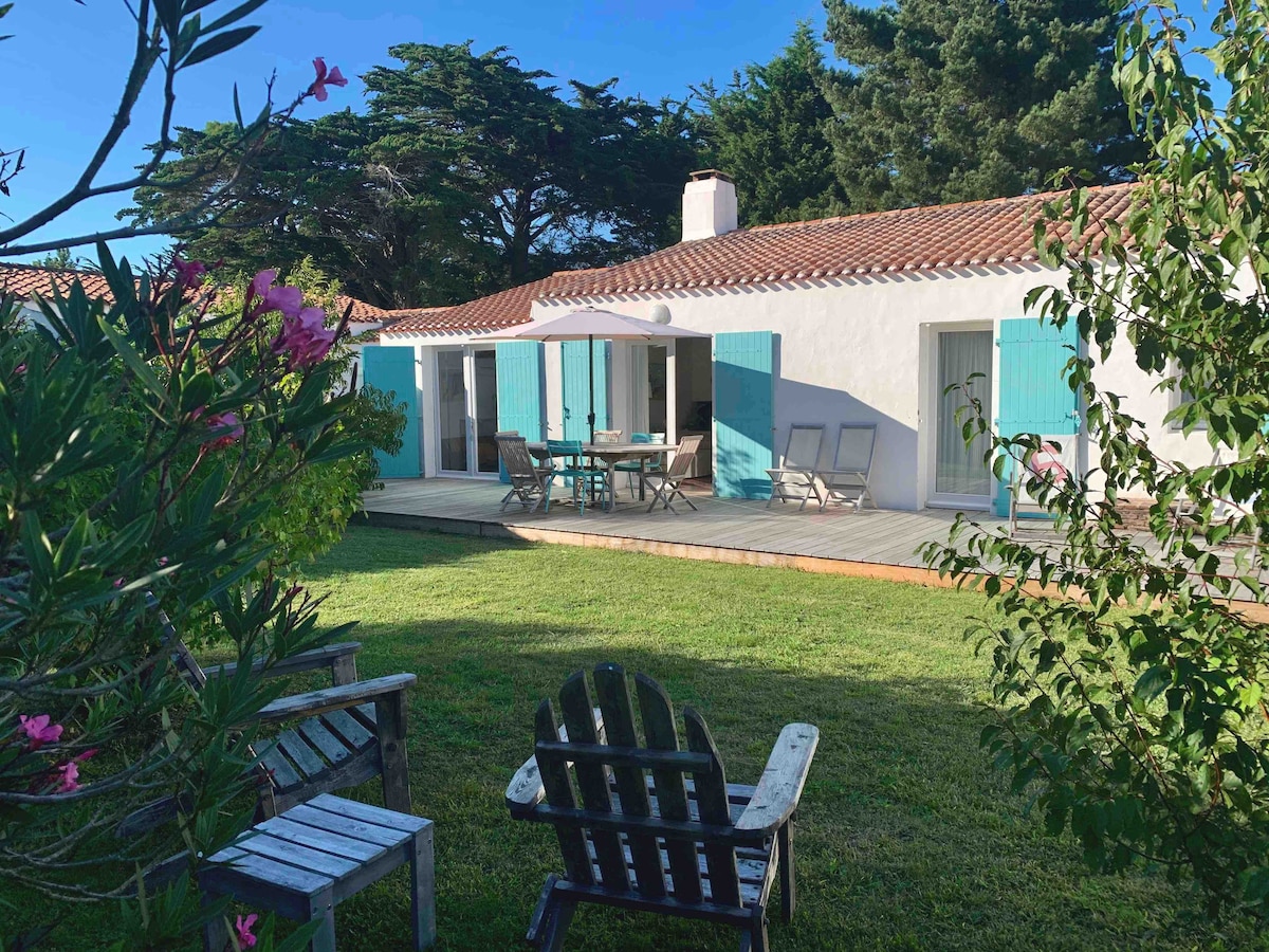 A charming house is visible with light blue shutters, surrounded by greenery. A spacious wooden deck features a dining table and chairs under a large umbrella, providing a shaded area for outdoor meals. Adjacent lounge chairs invite relaxation.