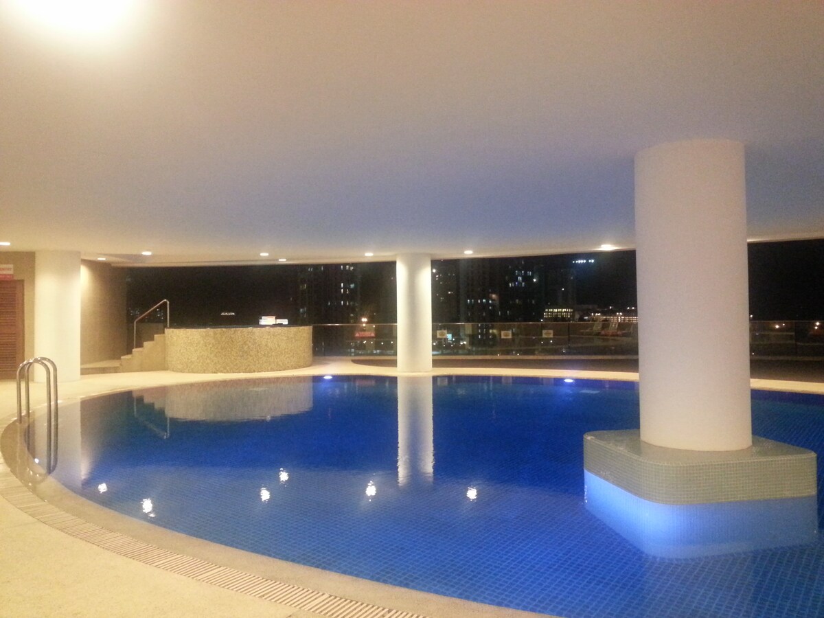 A well-lit indoor pool area features a spacious, blue-tiled pool with a circular design. Soft lighting reflects off the water's surface, creating a serene atmosphere. The area is framed by smooth walls and views of the outside city lights through large windows.