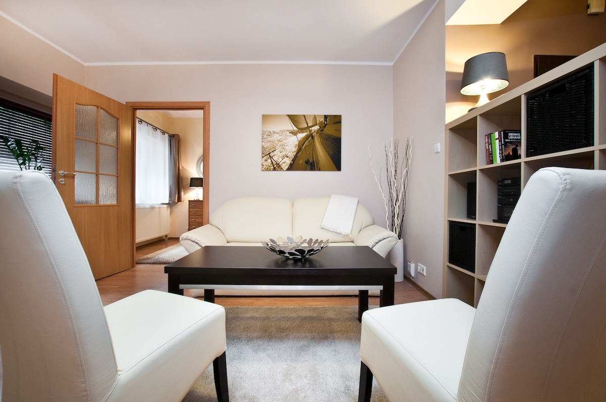 A contemporary living area is presented with a light-hued sectional sofa positioned in front of a dark coffee table. Two white armchairs flank the table, and a decorative piece is displayed atop it. A wooden shelf containing books is visible, along with a console in the background.