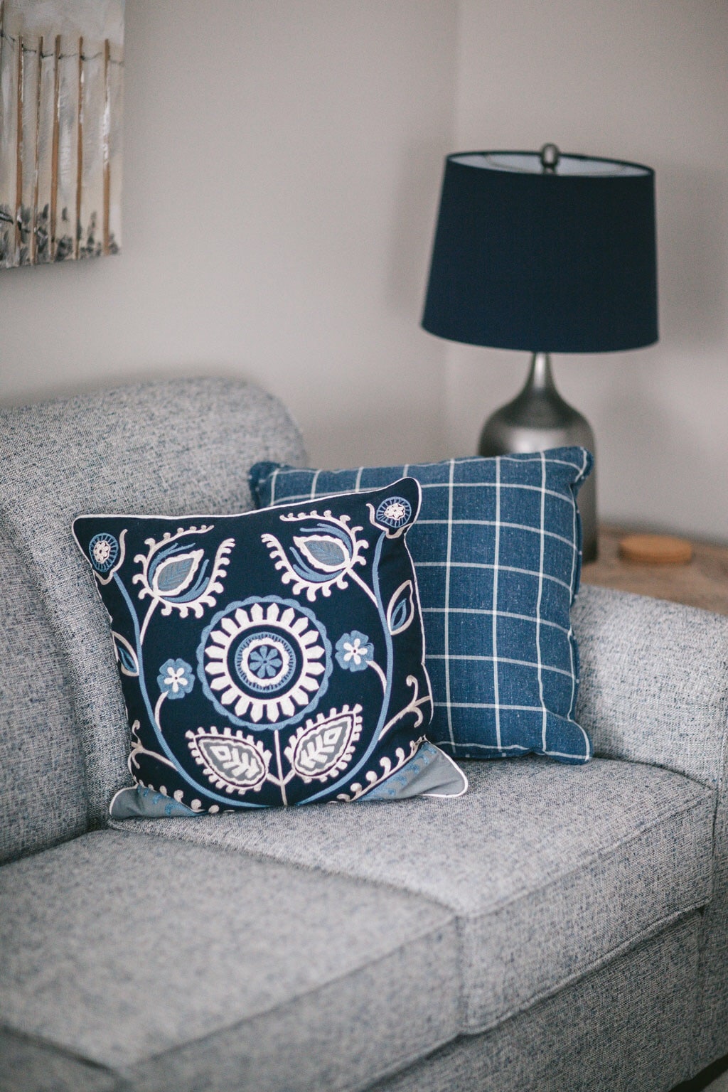 A gray upholstered sofa features two decorative pillows with intricate designs in shades of blue. A modern silver lamp with a navy shade is positioned beside the sofa, adding a subtle touch of elegance to the cozy corner.