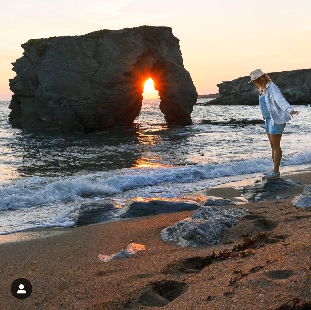 A golden sunset is framed by a natural rock arch along the shoreline. Gentle waves lap against the sandy beach, with scattered rocks and seaweed present. A person stands near the water's edge, dressed in light clothing, enjoying the serene coastal landscape.