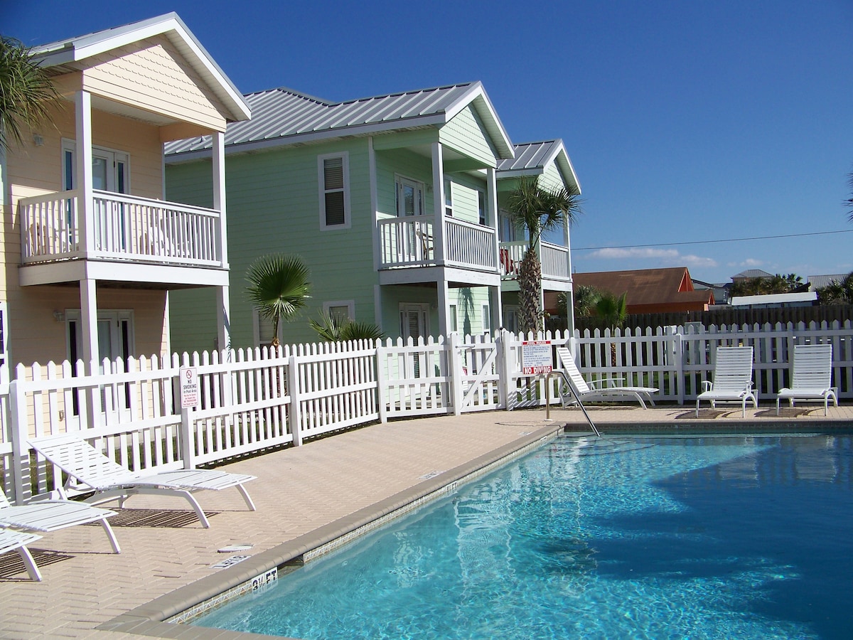 A pool area is shown with white lounge chairs arranged beside a clear blue pool. Two pastel-colored two-story buildings are visible in the background, framed by a white picket fence and palm trees, under a clear blue sky.