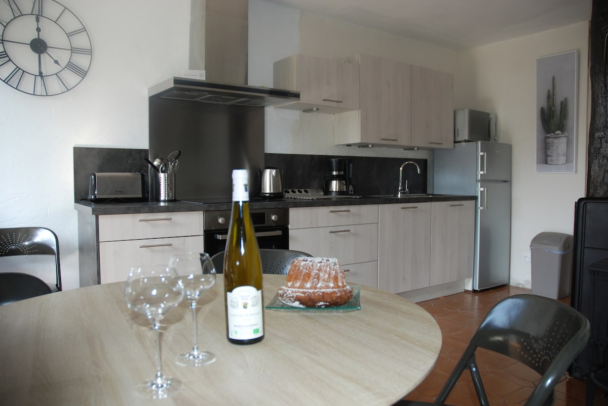 A modern kitchen area is presented, with wood-finished cabinetry and dark countertops. A round dining table is set with wine glasses and a bottle. A silver-colored refrigerator and various kitchen appliances are visible, contributing to a practical and welcoming cooking space.