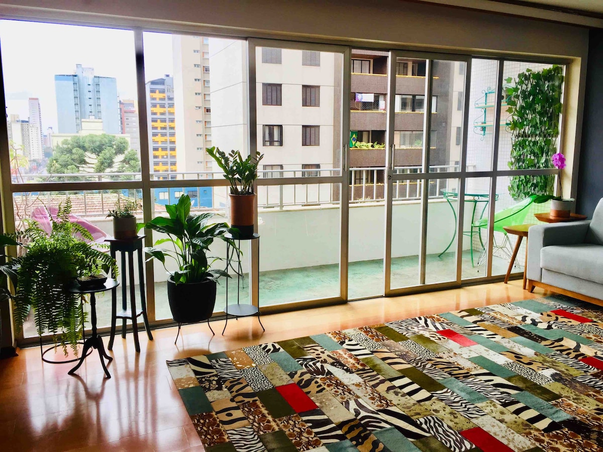 Large windows provide an urban view, allowing natural light to fill the space. Potted plants are arranged beside the windows, adding greenery. A colorful area rug with animal patterns covers the floor, complementing a comfortable seating area featuring a sofa.