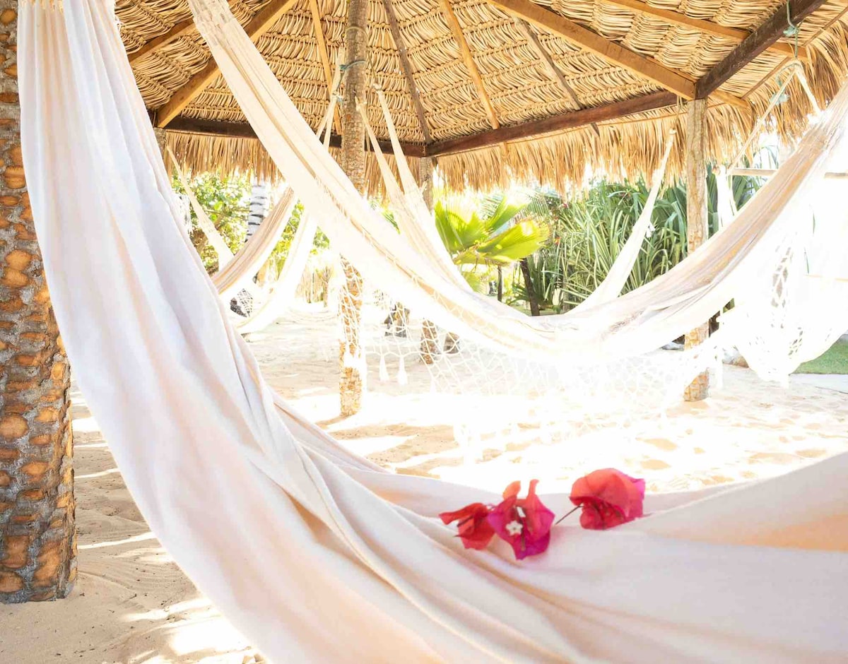 A shaded gazebo features multiple hammocks suspended from a thatched roof, creating a relaxing area surrounded by tropical greenery. Delicate flowers rest on one hammock, adding a touch of color to the neutral fabric. The sandy floor enhances the tranquil atmosphere.