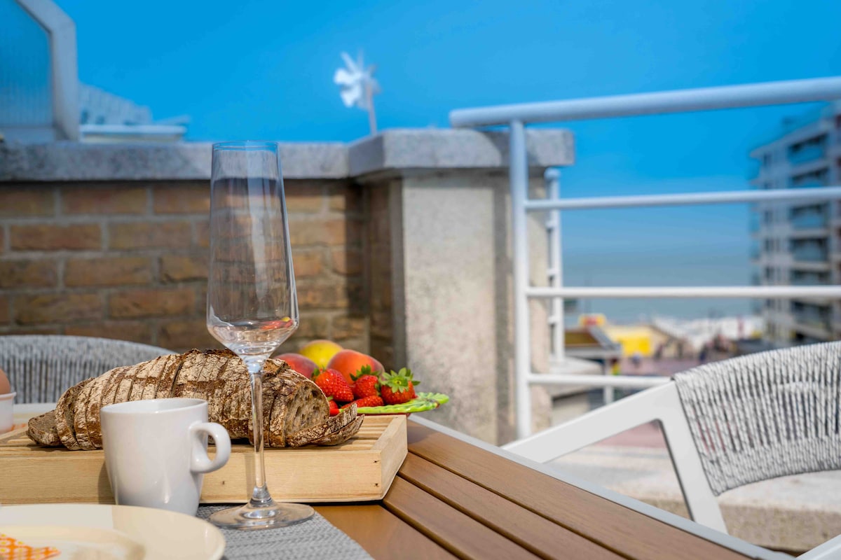A terrace table is set for breakfast with a wooden tray featuring fresh strawberries, croissants, and a glass of water. The ocean can be seen in the background, accompanied by modern buildings and a clear blue sky.