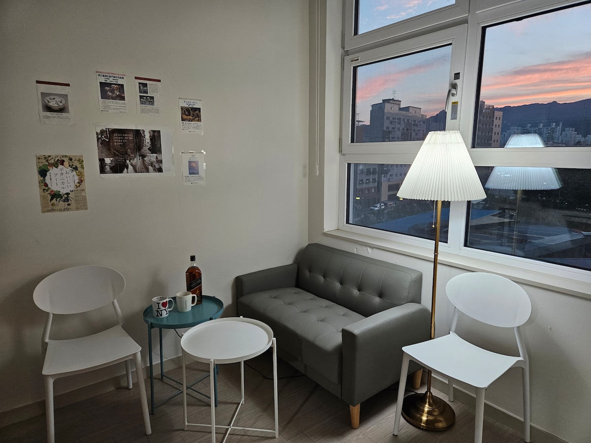 A cozy corner featuring a gray upholstered sofa paired with two white chairs. A glass table sits in the center, complemented by two coffee mugs. Soft light emanates from a stylish floor lamp, with a view of the city skyline visible through large windows.