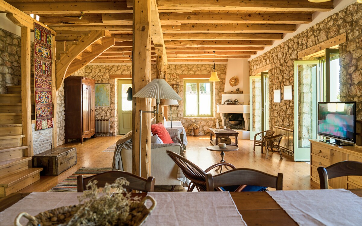 An open-concept living area is defined by wooden beams and stone walls. Natural light fills the space through large windows. A cozy seating arrangement with a television and a fireplace invite relaxation, while a dining table is visible in the foreground.
