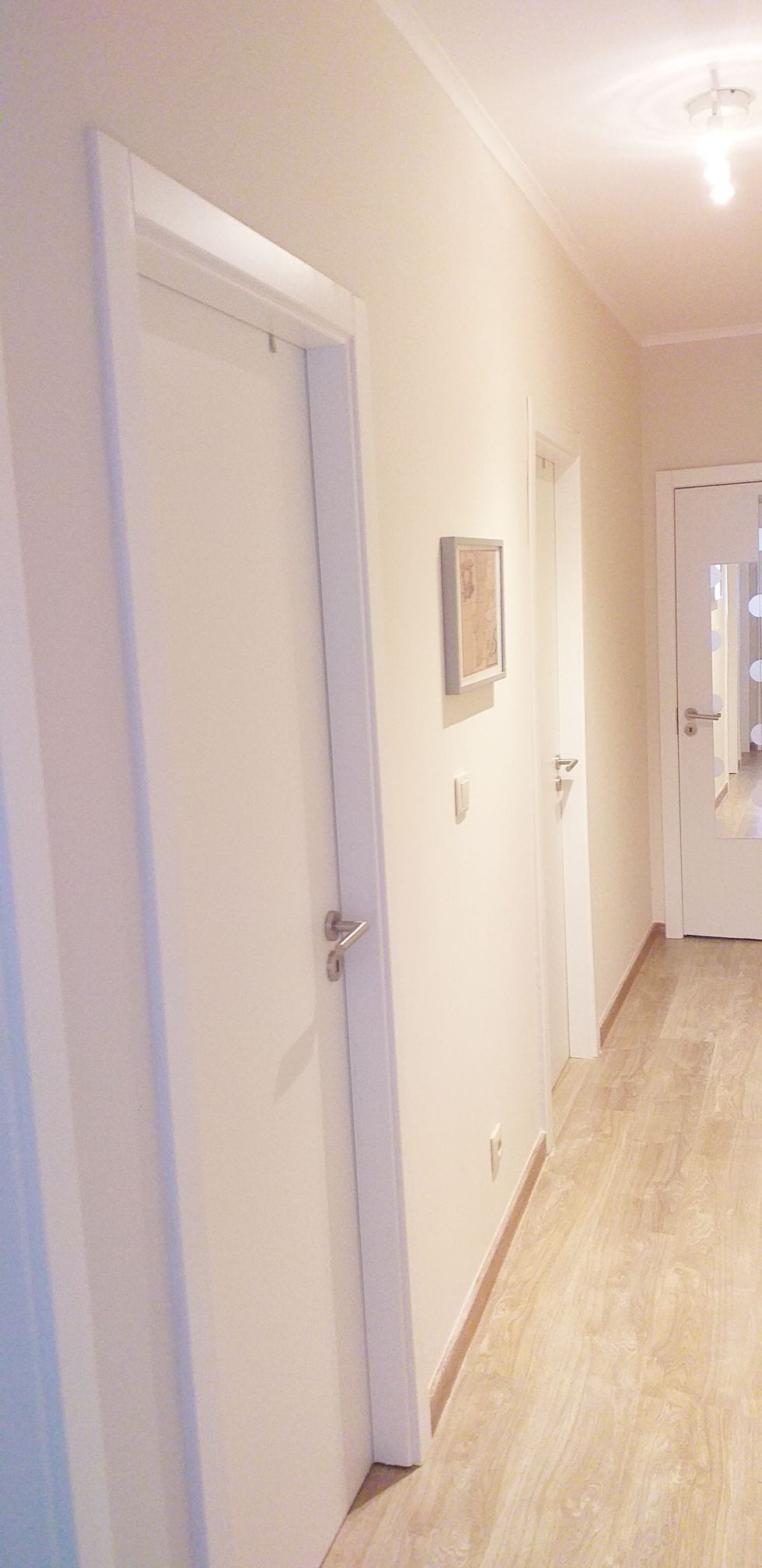 A well-lit hallway features light-colored walls and a smooth wooden floor. Two closed doors are visible, along with a mirror on a door at the far end. A framed picture hangs on the left wall, enhancing the corridor's simple and clean aesthetic.