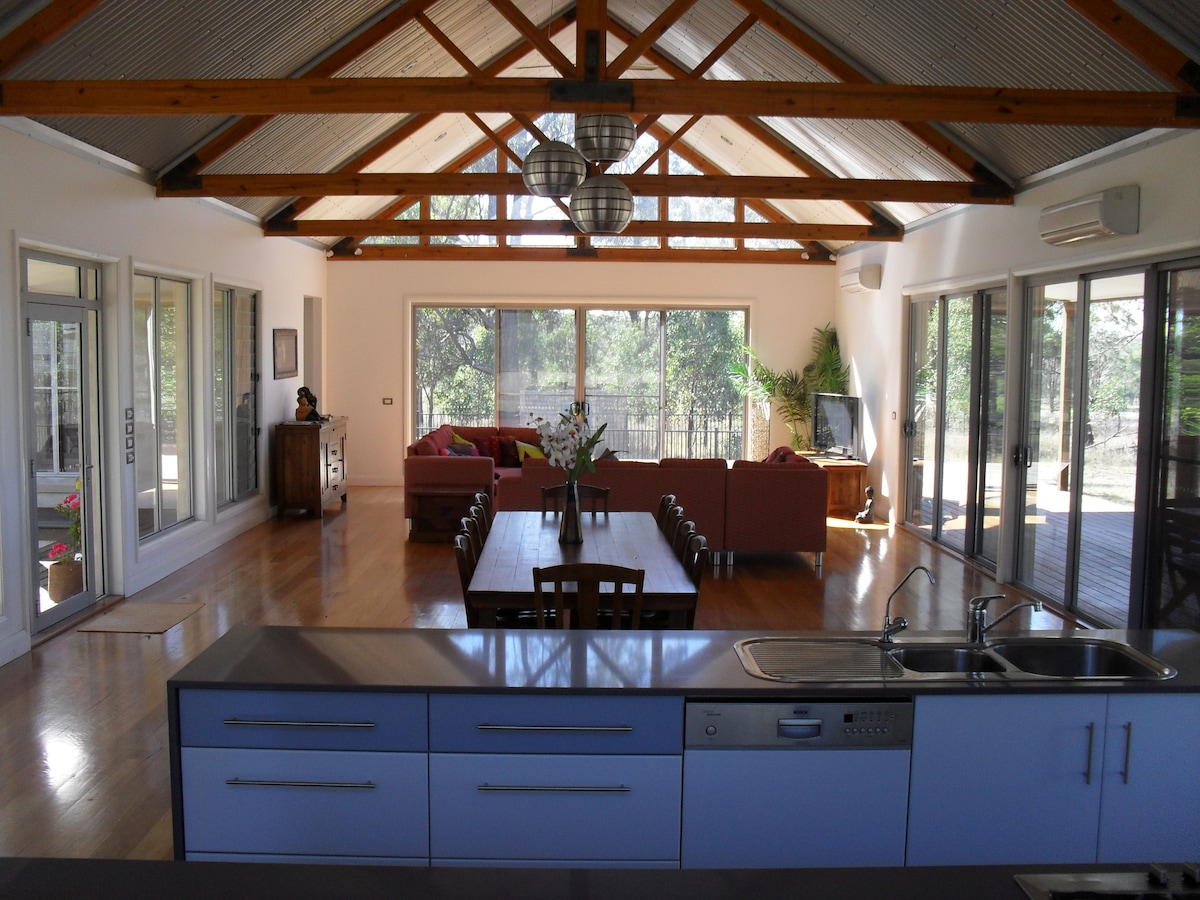 An airy open-plan living area showcases high wooden beams and large glass windows, providing ample natural light and views of the surrounding landscape. A spacious dining table is set, while a cozy sitting area with plush sofas is positioned nearby. The modern kitchen features a sleek countertop and appliances.