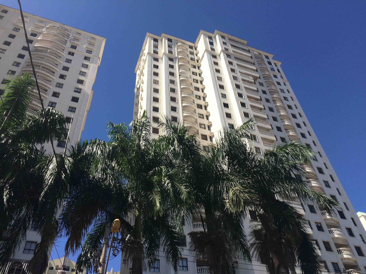 The building's modern architecture is showcased against a clear blue sky. Tall palm trees populate the foreground, adding greenery to the scene. The structure features multiple floors with prominent balconies and large windows, reflecting a bright, inviting environment.