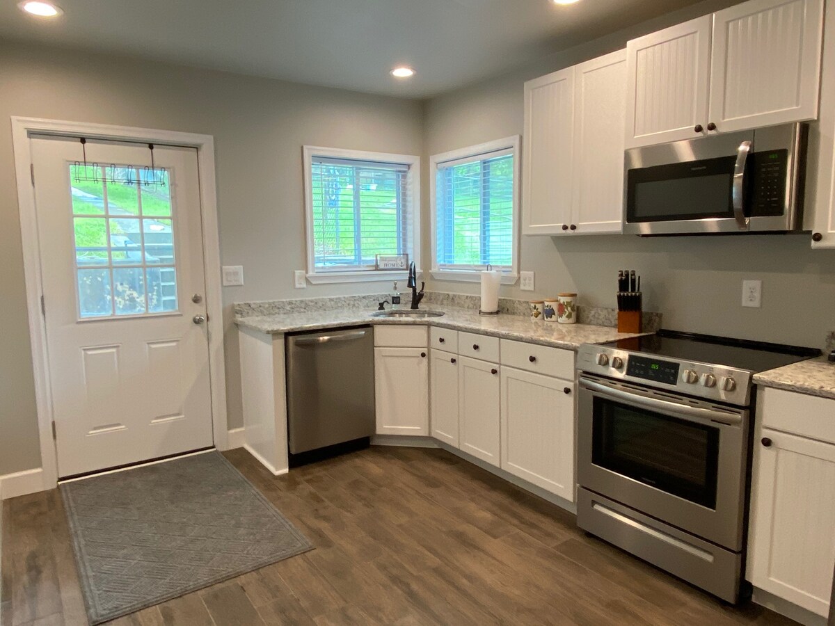 The kitchen features modern appliances including a stainless steel oven, microwave, and dishwasher. Granite countertops provide ample space for meal preparation. Natural light fills the room through two windows, enhancing the clean design with white cabinetry and a nearby door leading to the patio.