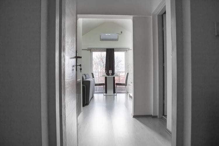 A minimalistic hallway leads to a living area with large windows allowing natural light. A small dining table with two chairs is positioned near the window, accompanied by a couch, creating a cozy space for relaxation.