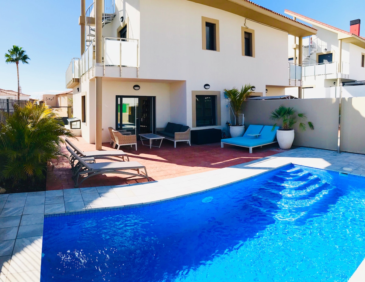 The outdoor area showcases a private heated pool with a clear blue surface, surrounded by loungers and seating arrangements. The villa features modern architecture, with large glass doors leading into the interior and palm trees enhancing the tropical ambiance.