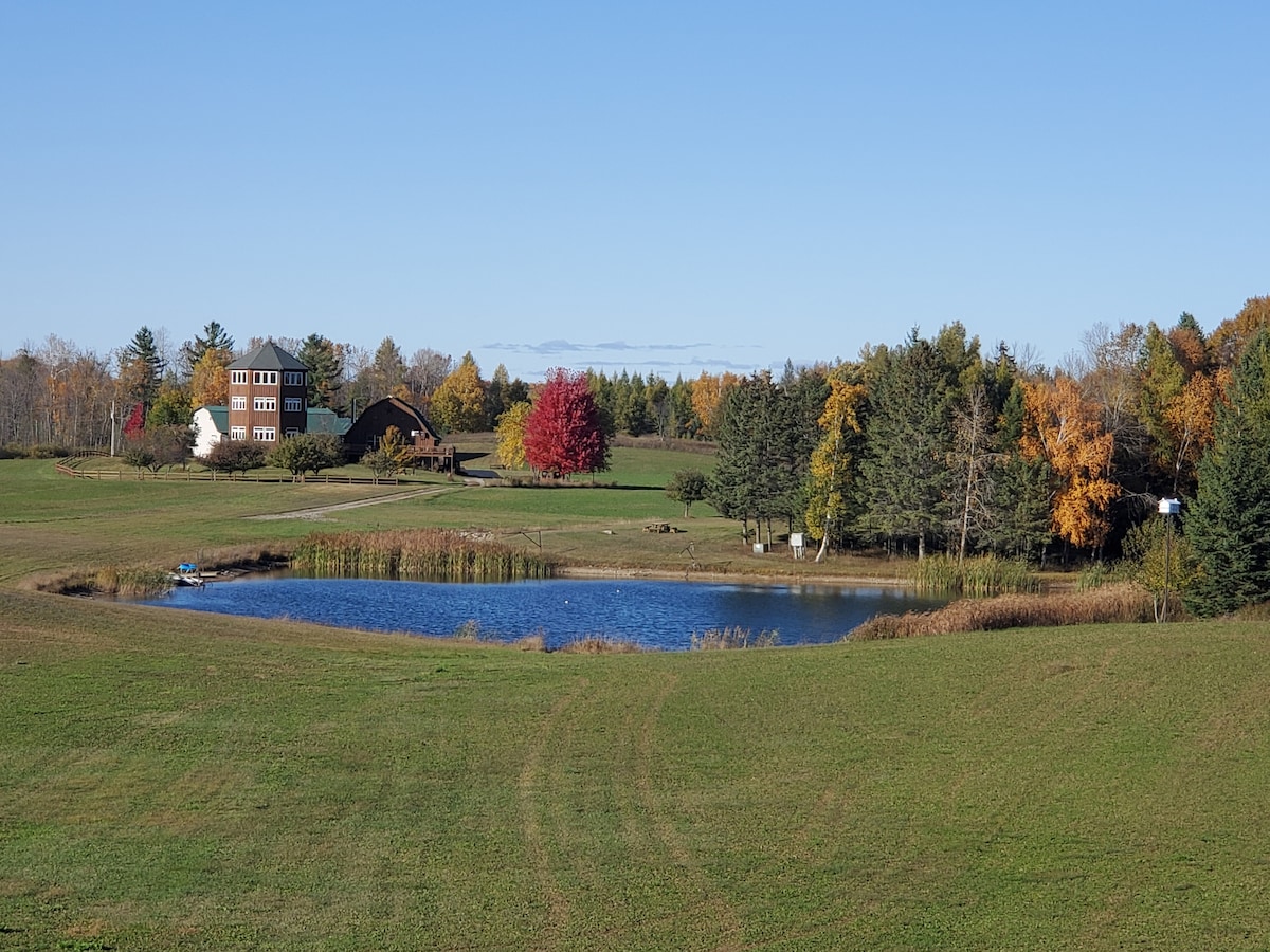 A serene landscape features a one-acre pond surrounded by lush greenery and colorful autumn foliage. A charming barn and the main lodge are visible in the distance, creating a harmonious blend with the natural setting.