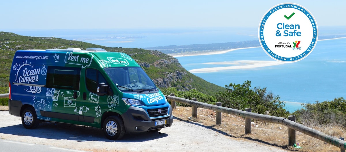 A campervan with a vibrant green and blue exterior is parked on a scenic overlook, framed by hills and the ocean in the distance. The coastline is visible, displaying sandy beaches and clear waters. A circular logo indicating cleanliness and safety is positioned in the corner.