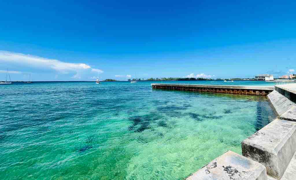 A clear view of the ocean waters showcases a blend of turquoise and aquamarine hues. The shoreline features a sturdy pier extending into the water, while sailboats can be seen in the distance against a backdrop of blue sky and scattered clouds.