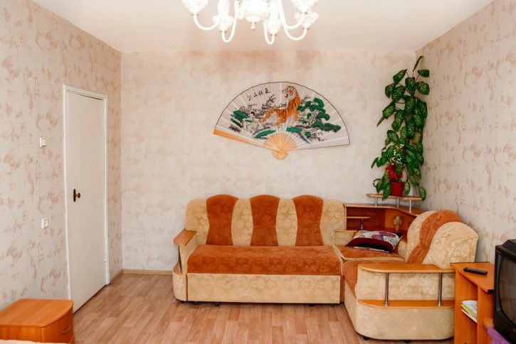 A living area features a comfortable two-tone sofa set in shades of orange and beige. A decorative wall fan depicting a tiger adds character. A small side table stands alongside the sofa, accompanied by a wooden unit and a solitary green plant in the corner.