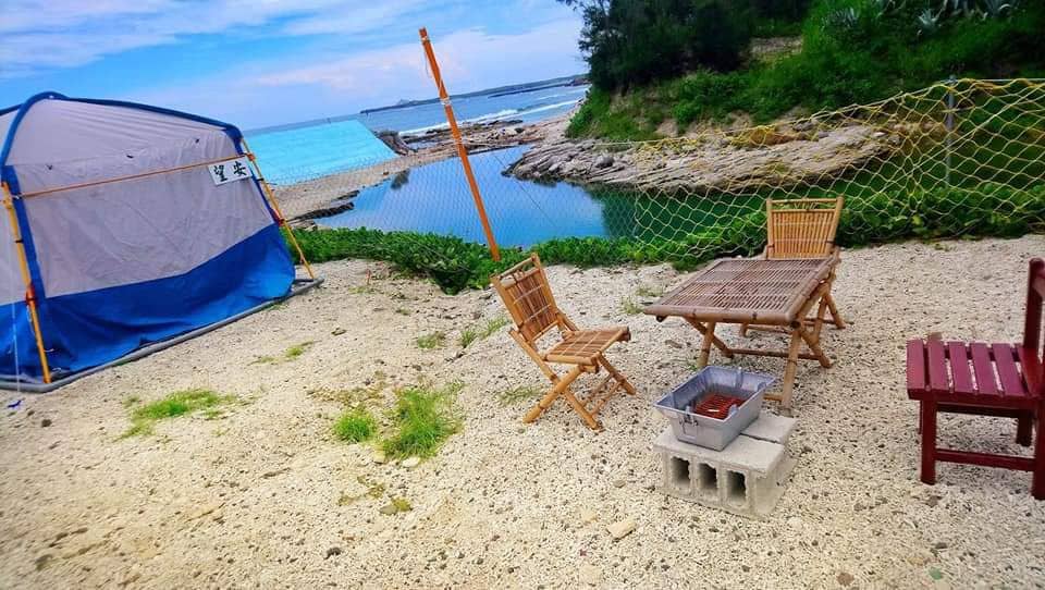 A camping area is featured beside a clear natural seawater pool. A tent is set up nearby, accompanied by wooden seating around a table. The ground is sandy, and a fire pit made from concrete blocks is positioned prominently, with the ocean visible in the background.