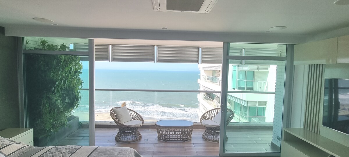 A cozy seating area features two woven chairs facing the ocean, complemented by a small round table. Expansive glass doors provide unobstructed views of the waves and sky, creating a bright and airy atmosphere within the penthouse.