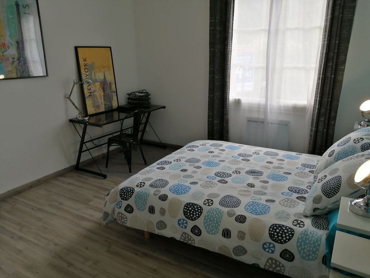 A bright sleeping area features a bed with a patterned comforter in shades of blue and gray. A streamlined desk and chair are positioned next to the window, where natural light filters through sheer curtains. Artwork is displayed on the adjacent wall.