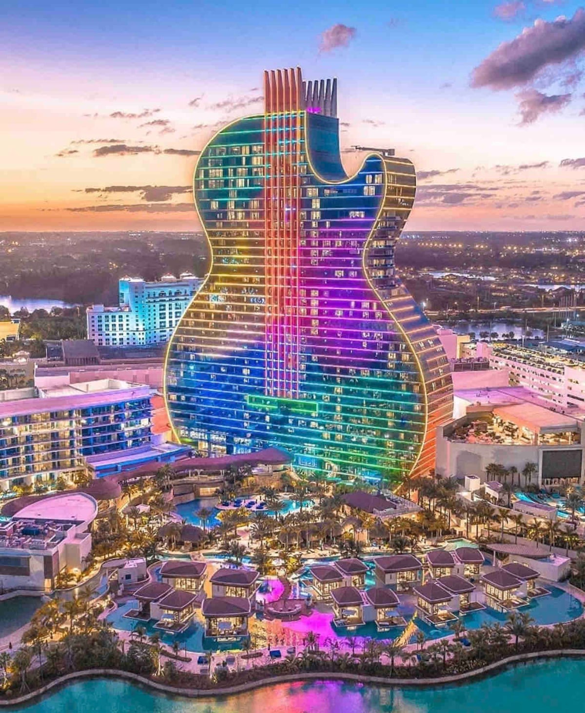 The iconic guitar-shaped hotel tower stands prominently against the twilight sky, featuring vibrant colors that illuminate its glass facade. Surrounding amenities include lush landscaping and a sparkling pool area, creating a lively atmosphere set in a resort-like environment.
