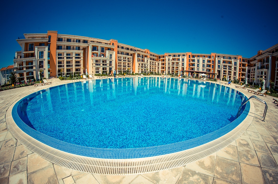 A large, circular swimming pool is framed by a spacious stone deck and lush landscaping. The pool's clear blue water reflects sunlight, while modern apartment buildings are visible in the background, showcasing a vibrant community atmosphere.