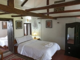 A well-appointed room features a spacious bed with crisp linens and a bedside lamp. Artistic wall decor adds character, while exposed wooden beams enhance the rustic charm. A wardrobe stands against the wall, providing ample storage, and a window allows soft natural light to enter.