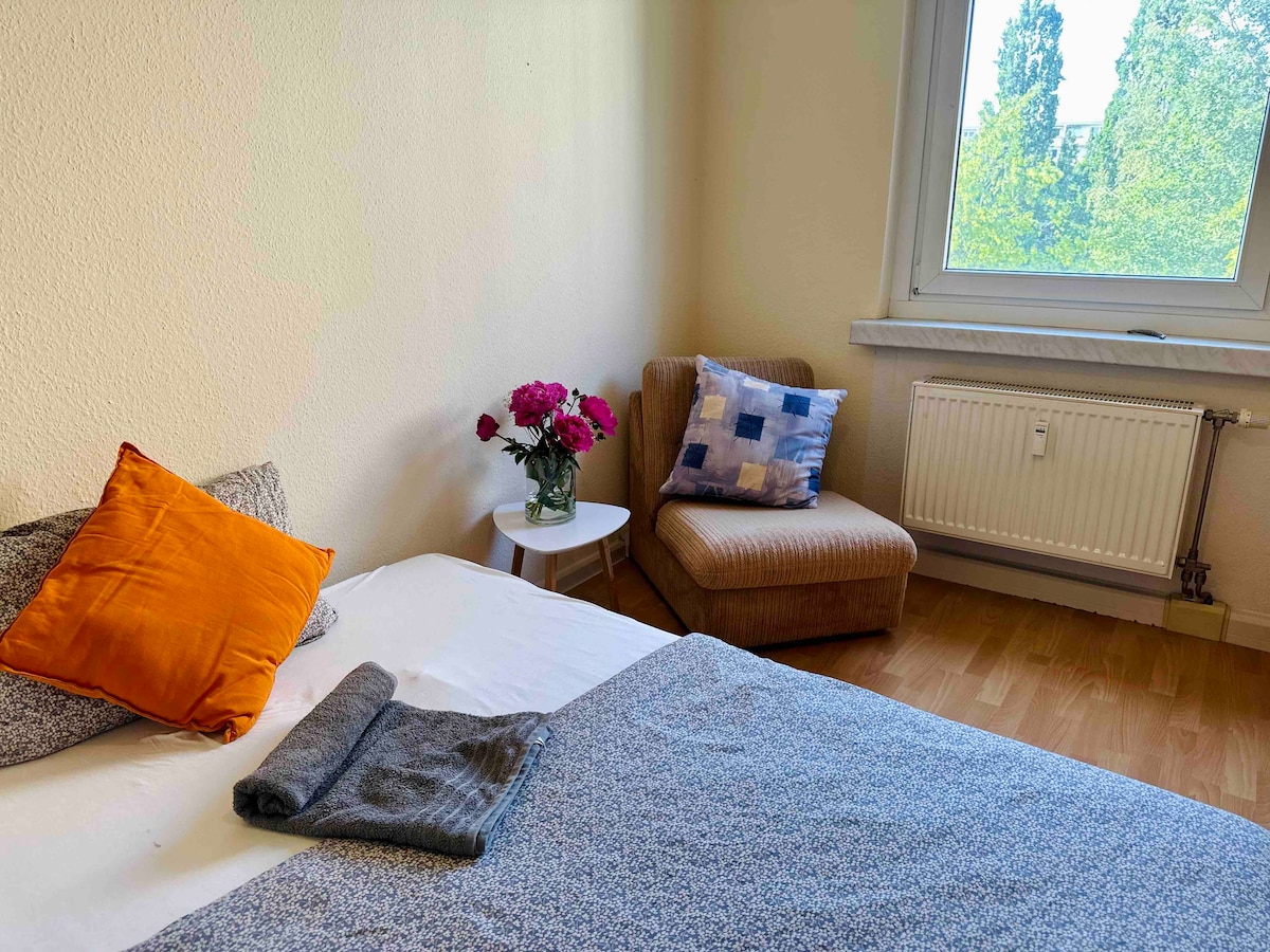 A simple yet comfortable bedroom is presented, featuring a spacious bed with decorative pillows and a folded blanket. A small side table holds a vase of fresh flowers. A cozy armchair is positioned beside the window, which offers natural light and a view of green trees outside.
