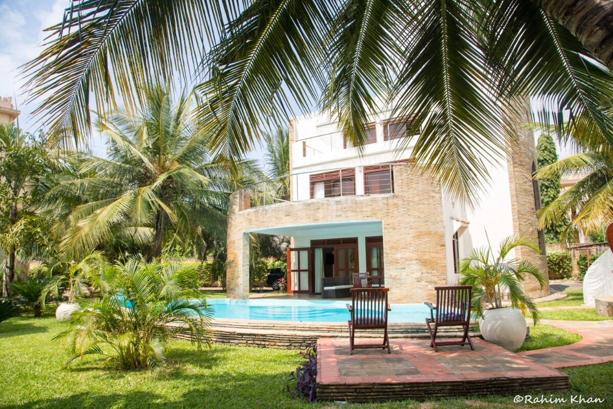 A modern two-story villa is surrounded by lush greenery, with palm trees framing the view. The property features a small swimming pool and is accentuated by a patio area furnished with two wooden chairs, inviting relaxation in the tranquil outdoor space.