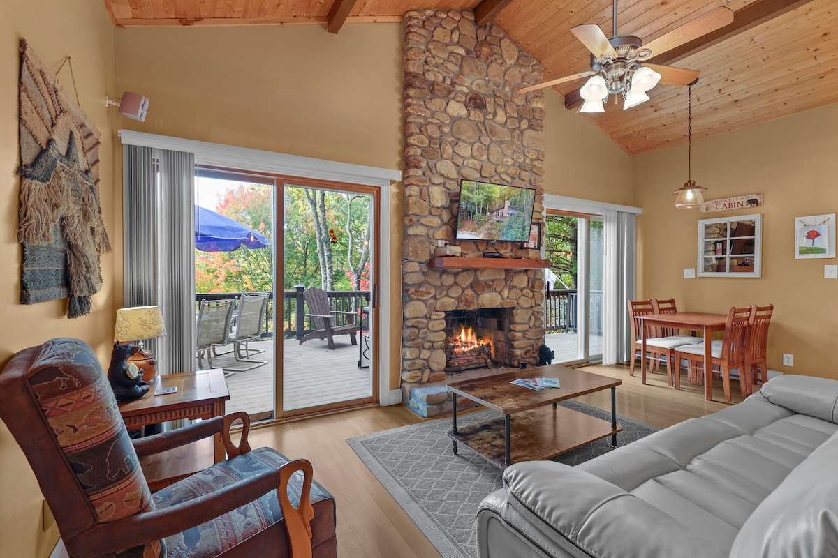 A comfortable living area displays a stone fireplace, contrasting with warm wooden beams overhead. Large glass doors provide views of an outdoor deck with seating. A cozy recliner and a medium-sized table with chairs are present, creating a welcoming space for relaxation and gathering.