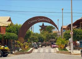 An archway marks the entrance to Canoa Quebrada, framed by greenery and a clear blue sky. The street is lined with rustic buildings, and vehicles are parked along the roadside, blending into the vibrant local scene.