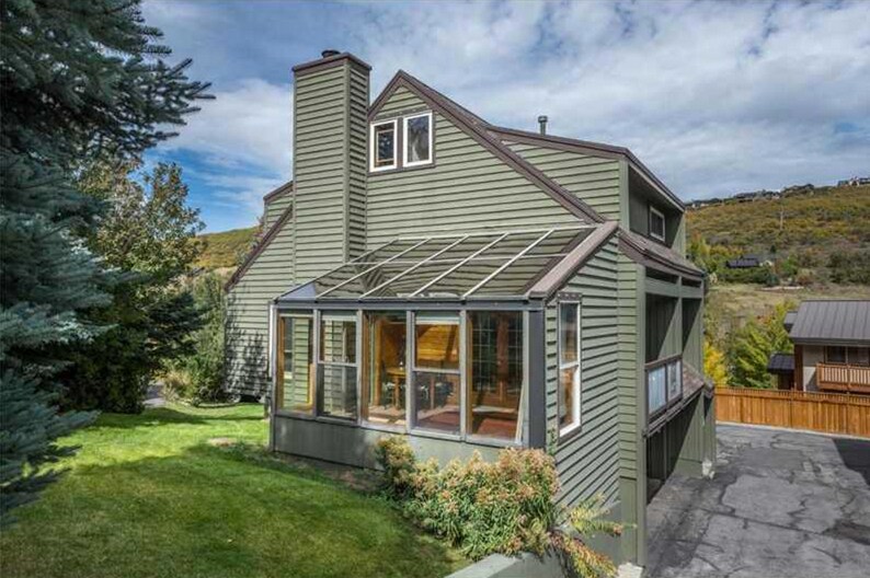 A green, multi-level townhouse is surrounded by landscaped grounds, featuring a glass-enclosed solarium attached to the side. Trees and shrubs provide natural greenery, contributing to the peaceful outdoor setting. The driveway is visible alongside the house, which showcases a well-maintained exterior.