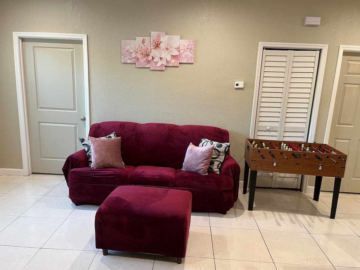 A comfortable burgundy sofa is positioned against a light-colored wall, adorned with decorative pillows. Above the sofa, floral artwork adds a subtle touch. A foosball table is situated nearby, while two doors lead to separate areas, enhancing the room's functionality.