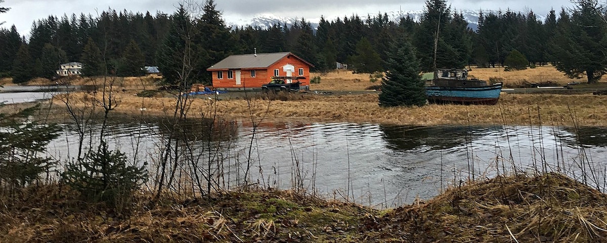 A vibrant orange cabin is positioned on the banks of a calm river, surrounded by tall green trees. The landscape features grassy patches along the water's edge and a distant blue boat on land, contributing to the natural scenery.