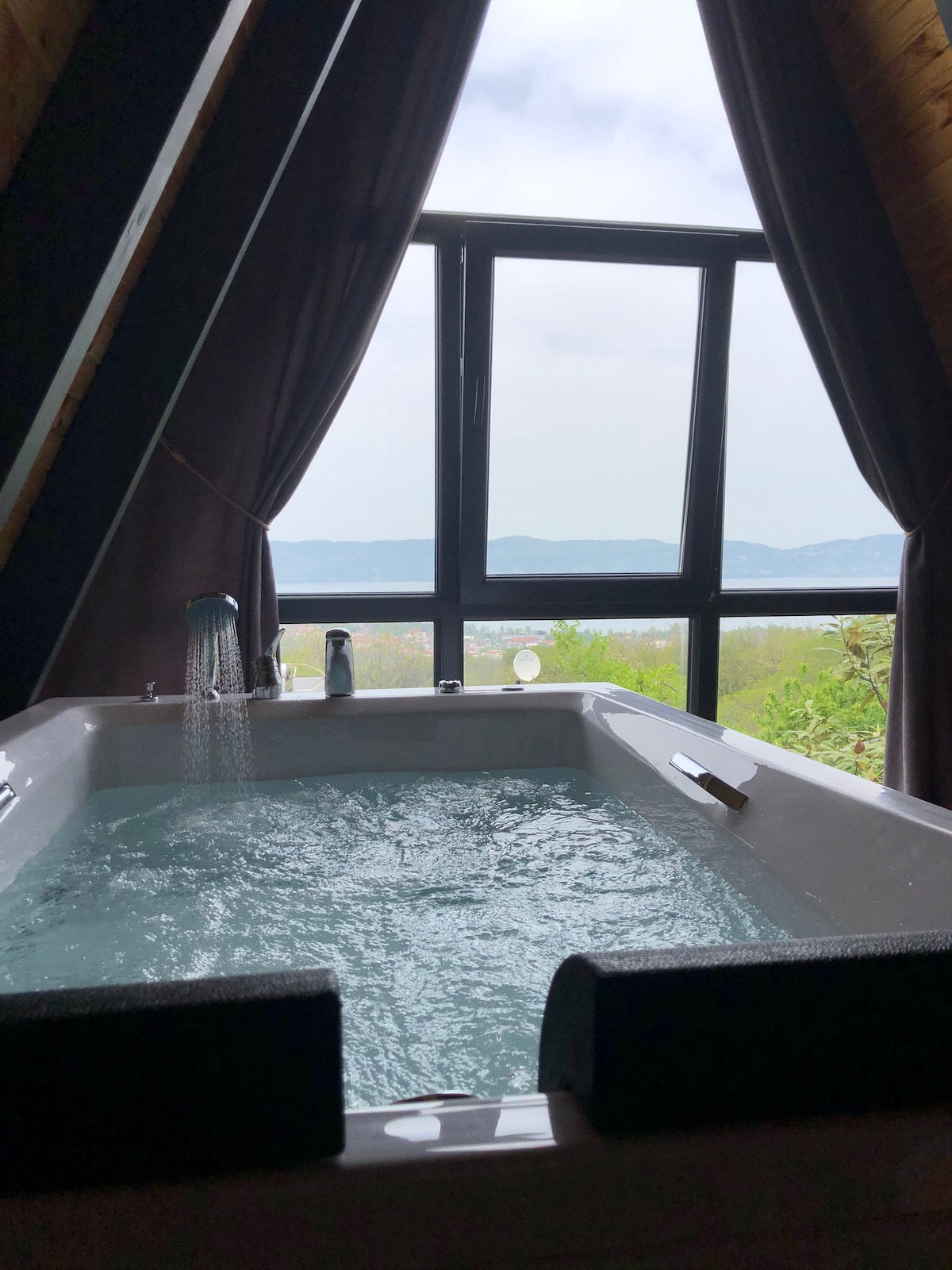 A spacious jacuzzi is positioned by a large window with a scenic view of the outdoors. Soft curtains frame the window, allowing natural light to illuminate the bubbling water. Green hills can be seen in the distance, creating a serene backdrop.